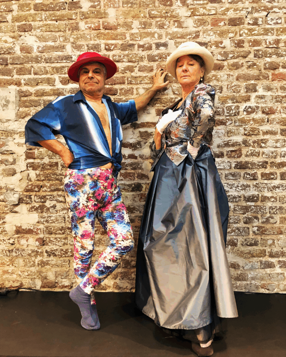 Zwei ältere Personen stehen vor einer Backsteinwand. Die linke Person trägt einen roten Hut, ein blaue-weißes, offenes und an der Taille geknotetes Hemd und eine bunt gemusterte Hose. Die rechte Person trägt eine beigefarbene Hut, weiße Handschuhe, ein silbrig glänzendes, geblümtes Oberteil und einen langen grauen Rock.