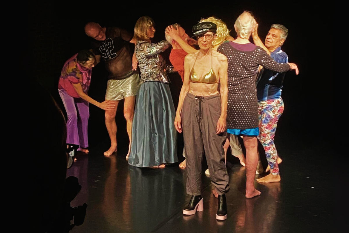 Eine Gruppe älterer Menschen auf einer Bühne, die bunte, auffällige Kleidung trägt. Im Vordergrund steht eine ältere Frau, die einen goldenen BH trägt.