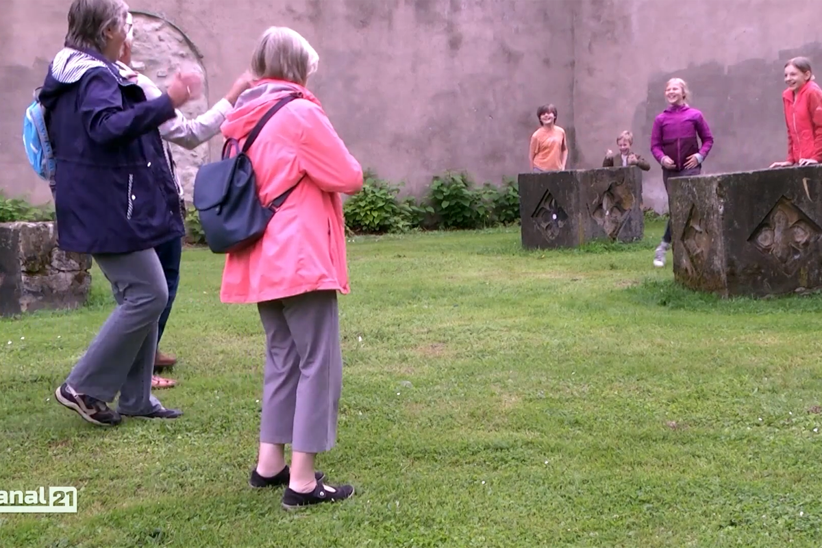 Mehrere ältere Personen stehen auf einer Wiese vor einer hohen Steinmauer. Vor der Mauer liegen quaderförmige Steine mit ornamentalen Mustern, hinter denen Kinder stehen.