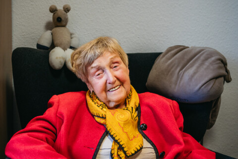 Eine ältere Frau sitzt lächelnd in einem Sessel. sie trägt eine rote Jacke und ein gelbes Halstuch. Auf der Sessellehne sitzt ein Stofftier.