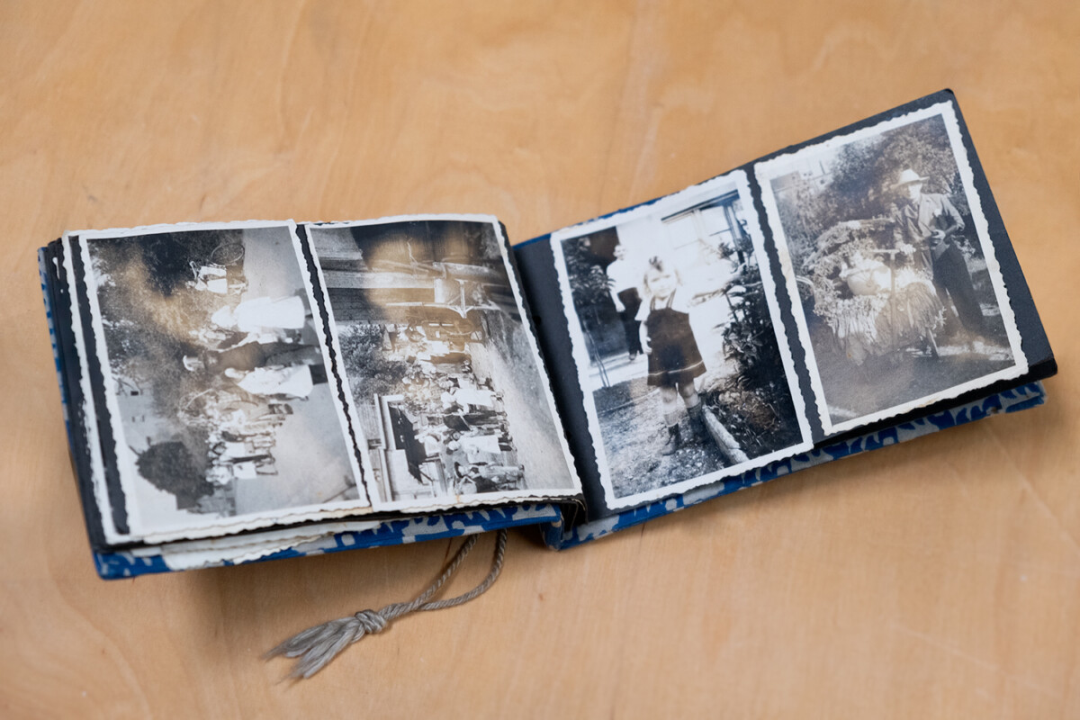 Ein aufgeschlagenes, altes Fotoalbum liegt auf einem Holztisch und zeigt vier Schwarz-Weiß-Fotografien. Die Bilder zeigen Szenen aus vergangenen Zeiten, darunter eine Gruppe von Menschen im Freien, ein kleines Mädchen im Kleid sowie einen älteren Mann neben einem Kinderwagen oder geschmückten Wagen im Garten.