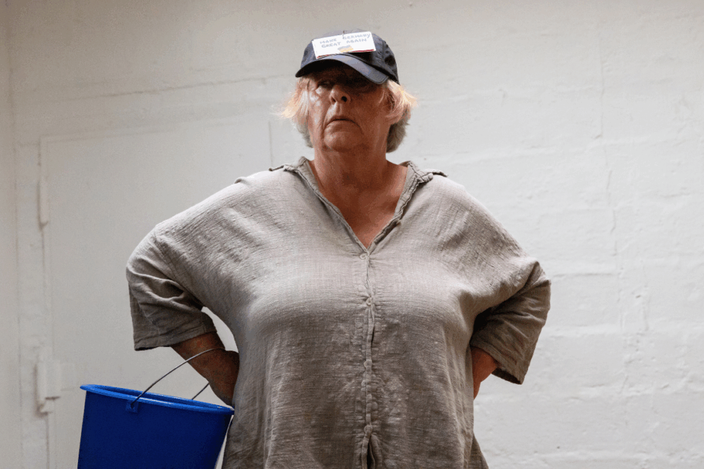 Eine ältere, weiblich gelesene Person trägt eine Kappe, auf der ein Zettel mit der Aufschrift „Make Germany great again“ angebracht ist. Ihre Hände sind in die Hüften gestemmt. Am rechten Arm hängt ein blauer Eimer,