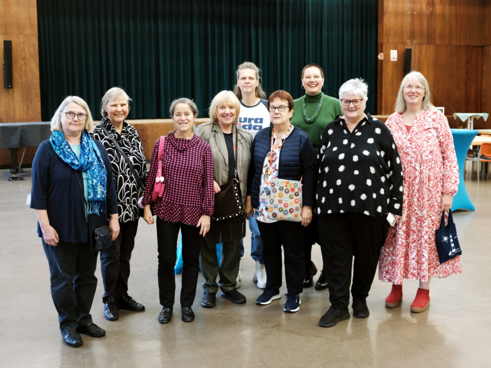 Gruppenfoto mit neun überwiegend ältere Frauen in einem Saal vor einer Bühne mit dunklem Vorhang. Sie tragen gemusterte Kleidung und Taschen.