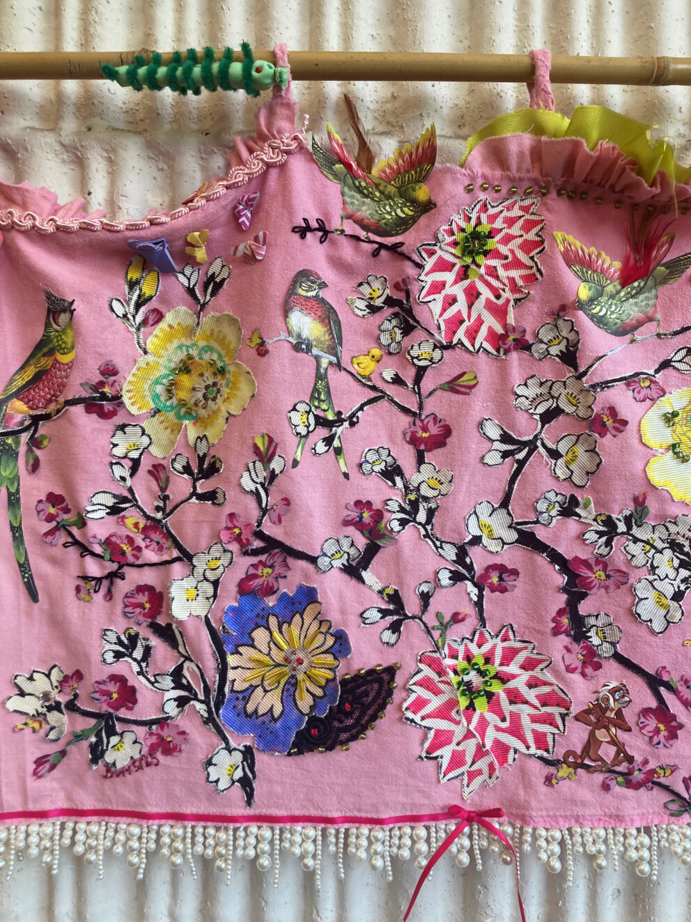 Ein rosa Stoffstück hängt an einer Stange. Darauf sind bunte Blumen und Vögel appliziert. Am unteren Rand ist eine Bordüre aus weißen Perlen befestigt.
