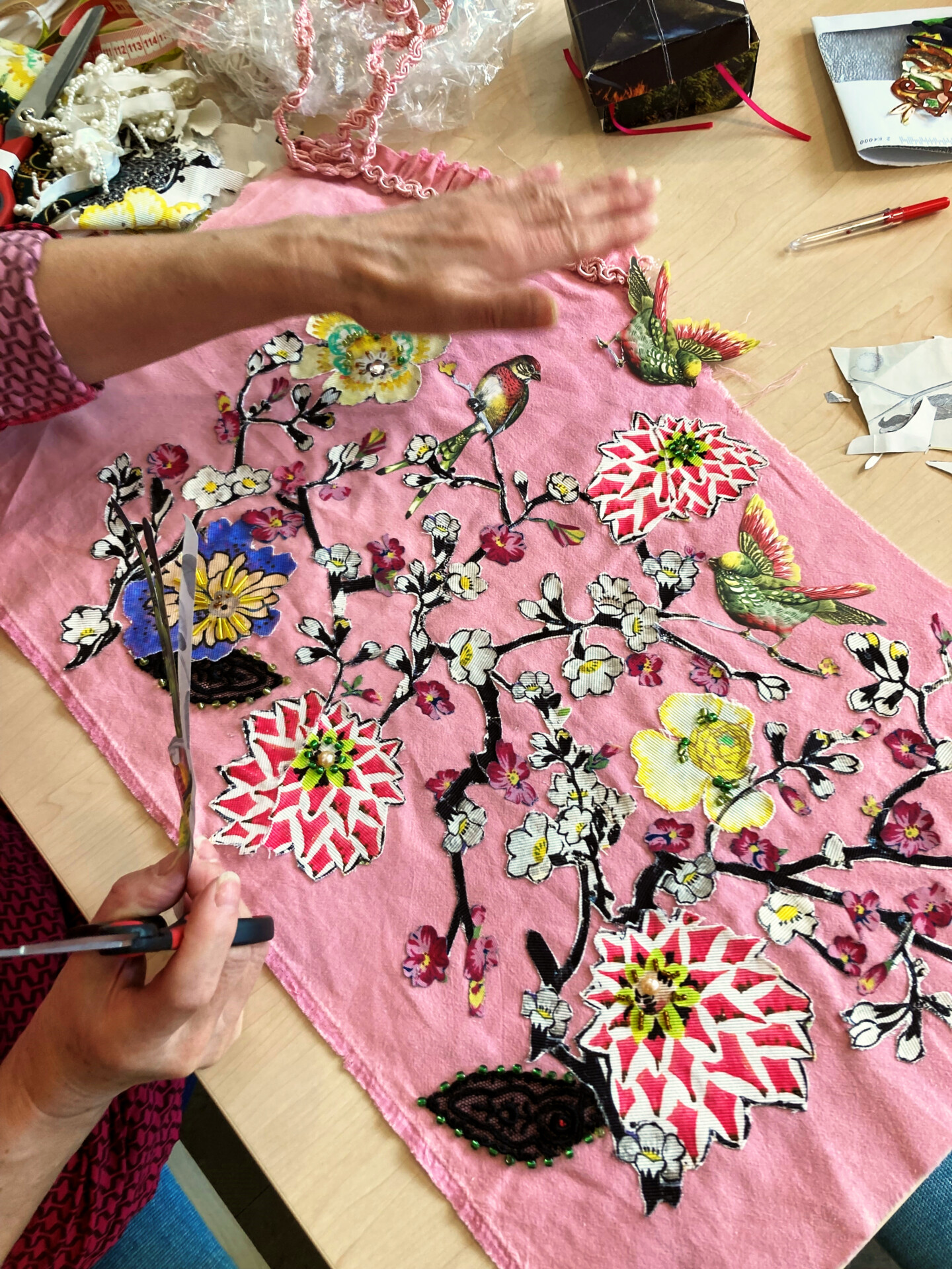 Ein rosa Stoffstück liegt auf einem Tisch. Darauf sind bunte Blumen und Vögel appliziert. Eine Hand hält eine Schere, daneben liegen Perlen andere Materialien.