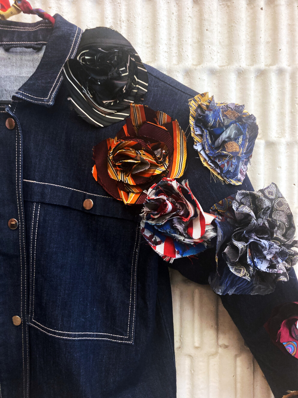 Eine dunkle Jeansjacke, an deren Schulter mehrere große Stoffblüten aus bunten, gemusterten Materialien befestigt sind.