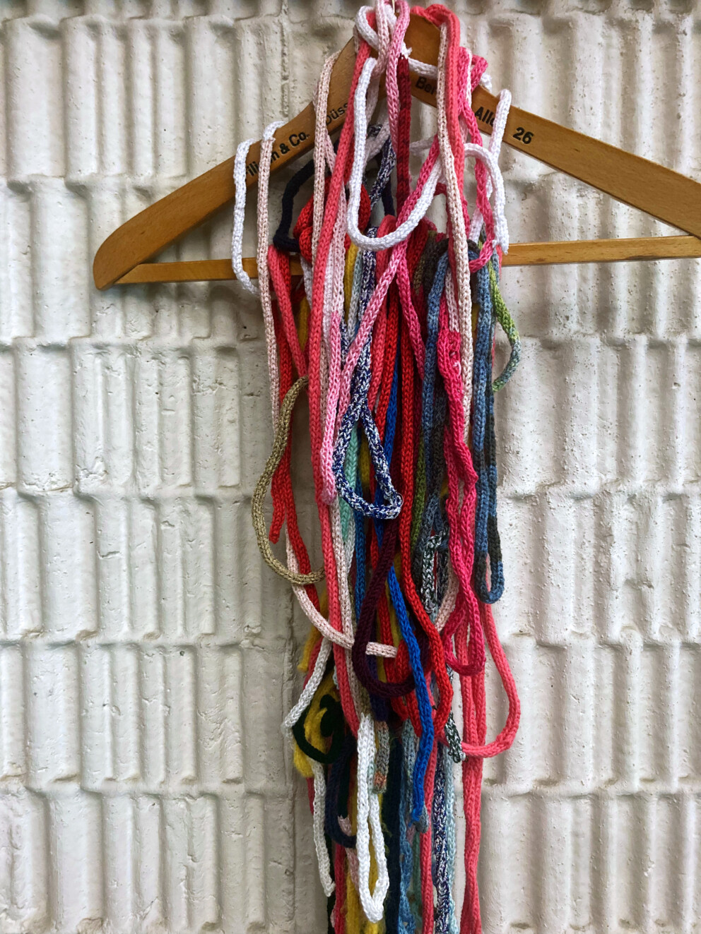 Ein Kleiderbügel hängt an einer Wand. Daran hängen viele bunte, gestrickte Schnüre in verschiedenen Farben wie Pink, Blau, Grün und Weiß.
