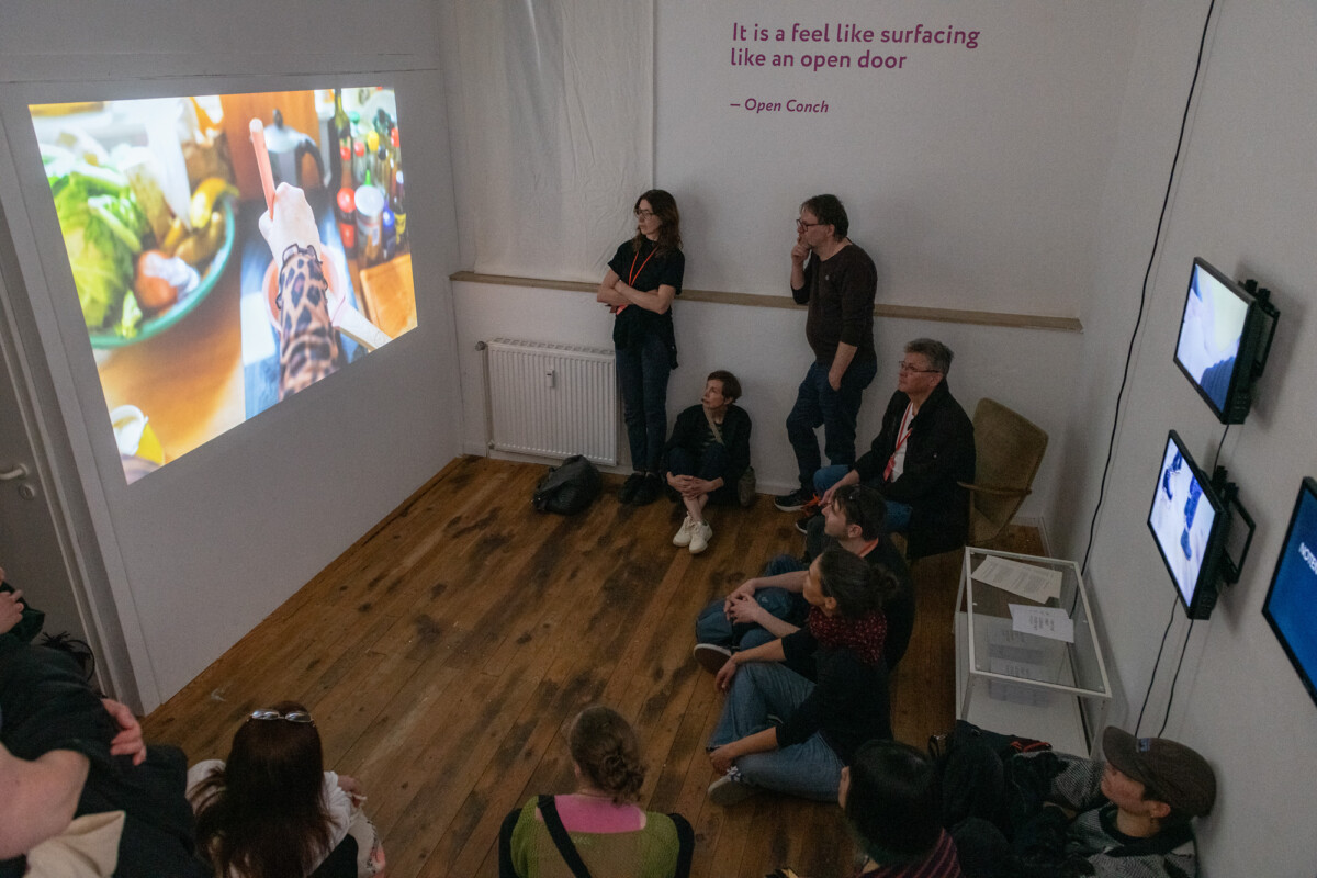 Mehrere Personen sitzen und stehen in einem kleinen Raum mit Holzfußboden. An der linken Wand wird ein Video mit einer Küchenszene projiziert. Rechts hängen drei Bildschirme. An der Wand steht der Text: „It is a feel like surfacing like an open door – Open Conch“.