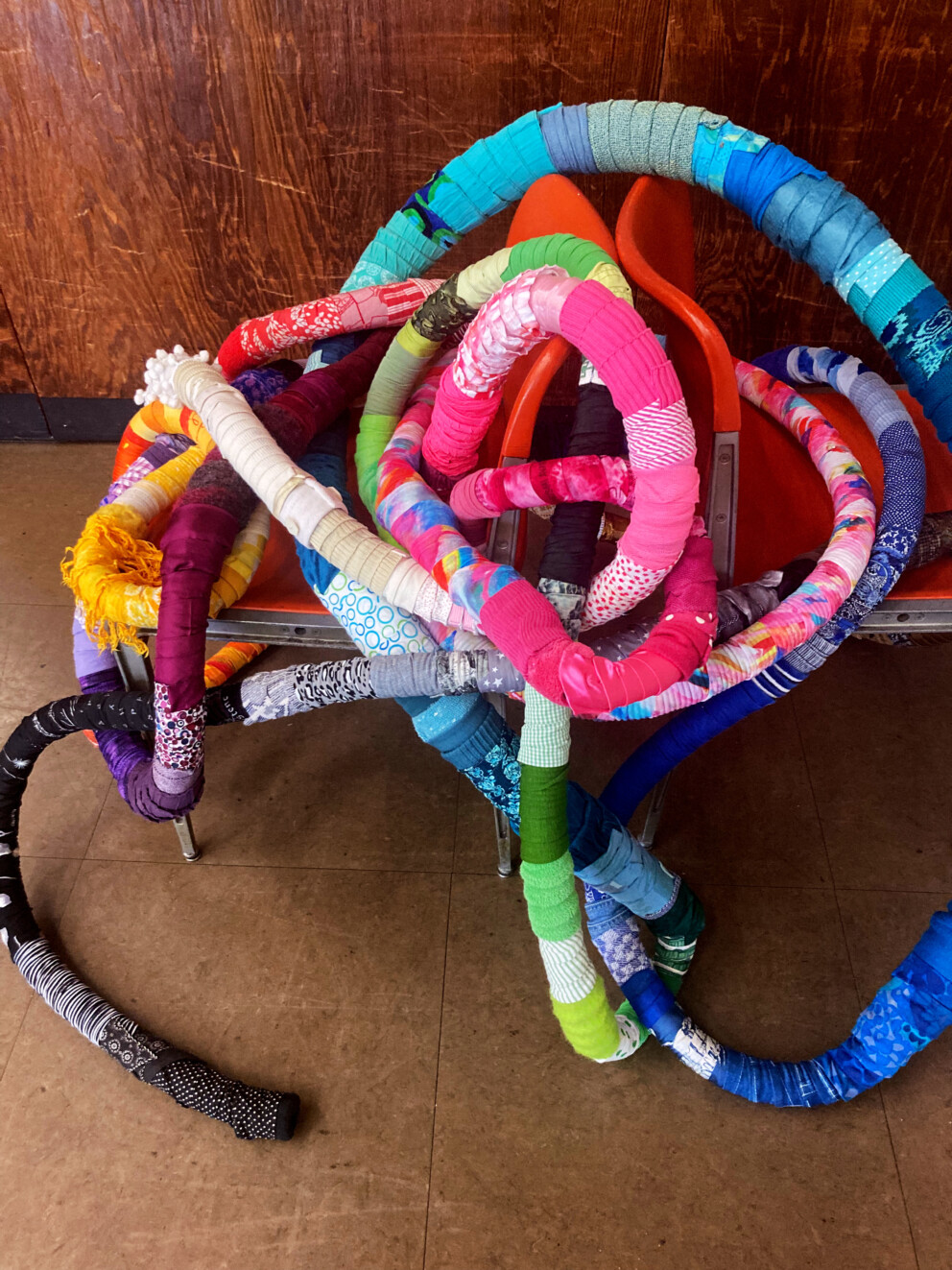 Mehrere lange, dicke Stoffschläuche in bunten Farben liegen verschlungen auf roten Stühlen. Die Stoffe sind gemustert und in kräftigen Farbtönen.