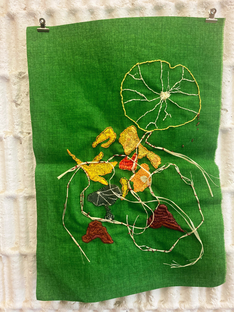 Ein grünes Stoffstück hängt an einer Wand. Darauf sind gestickte Formen in Gelb, Braun und Rot sowie lose Fäden und Garnstücke.