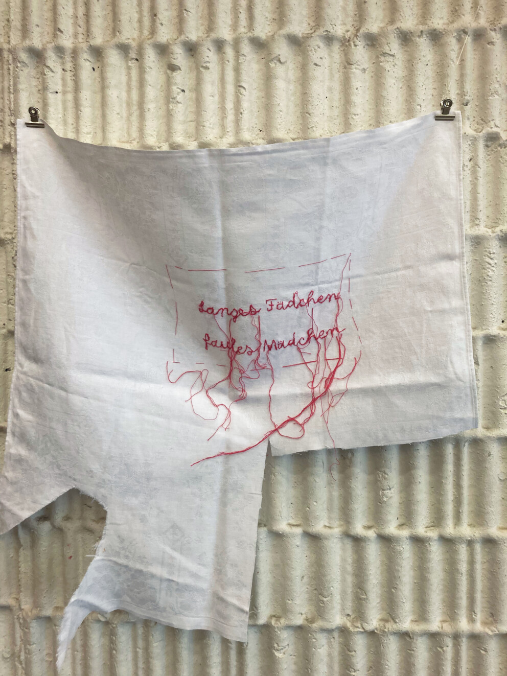 Ein weißes Stofftuch hängt an einer Wand. Darauf ist eine rote Stickerei mit lose hängenden Fäden zu sehen und Schriftzug in Schreibschrift: „Langes Fädchen, faules Mädchen“.