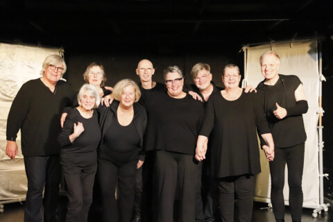 Eine Gruppe von neun älteren Erwachsenen steht auf einer Bühne. Alle tragen schwarze Kleidung und stehen eng zusammen, einige haben die Arme umeinander gelegt oder halten sich an den Händen. Hinter ihnen stehen zwei mit hellem Stoff bespannte Bühnenelemente.