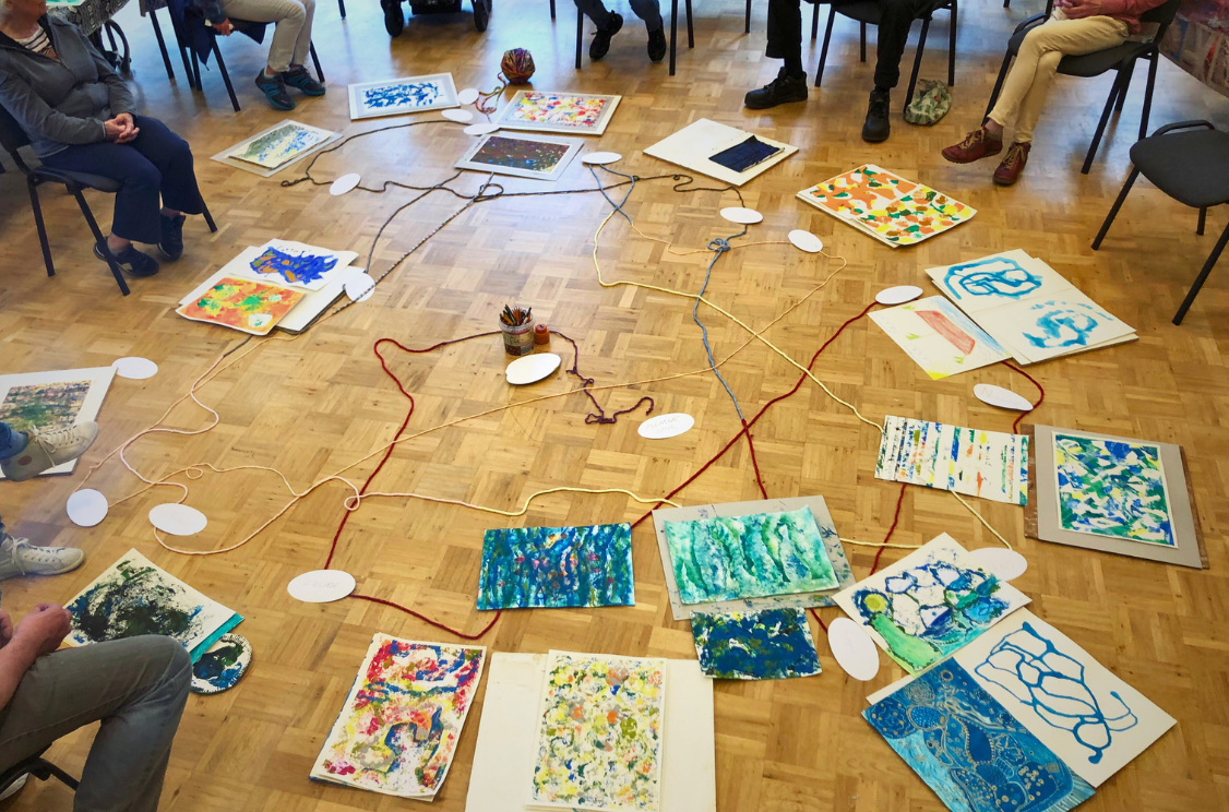 Eine Gruppe von Menschen sitzt in einem Kreis auf Stühlen in einem Raum mit Holzboden. In der Mitte des Kreises liegen verschiedene bunte Kunstwerke und Zeichnungen auf dem Boden verteilt. Mehrere Seile und Schnüre verbinden die Kunstwerke miteinander, und einige ovale Papierstücke sind ebenfalls auf dem Boden verteilt.