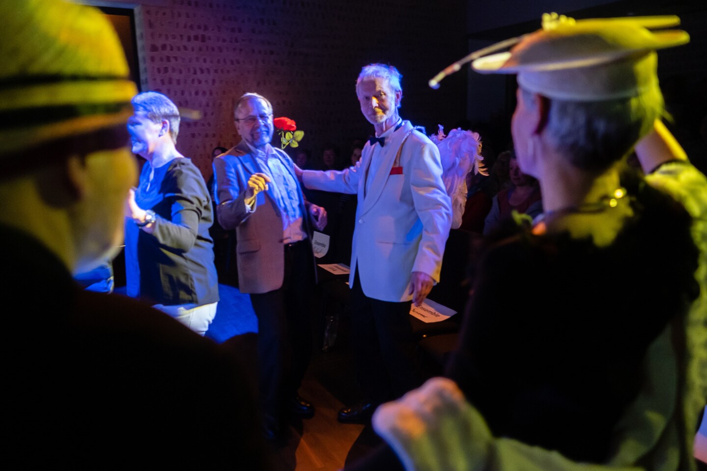 Im Zentrum der Fotografie stehen zwei ältere Männern in schicker Kleidung. Einer hat eine Rose in der Hand.