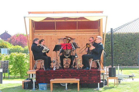 auf einer Wiese steht eine kleine Holzbühne. Auf der Holzbühne sitzen 5 Personen und spielen Instrumente.