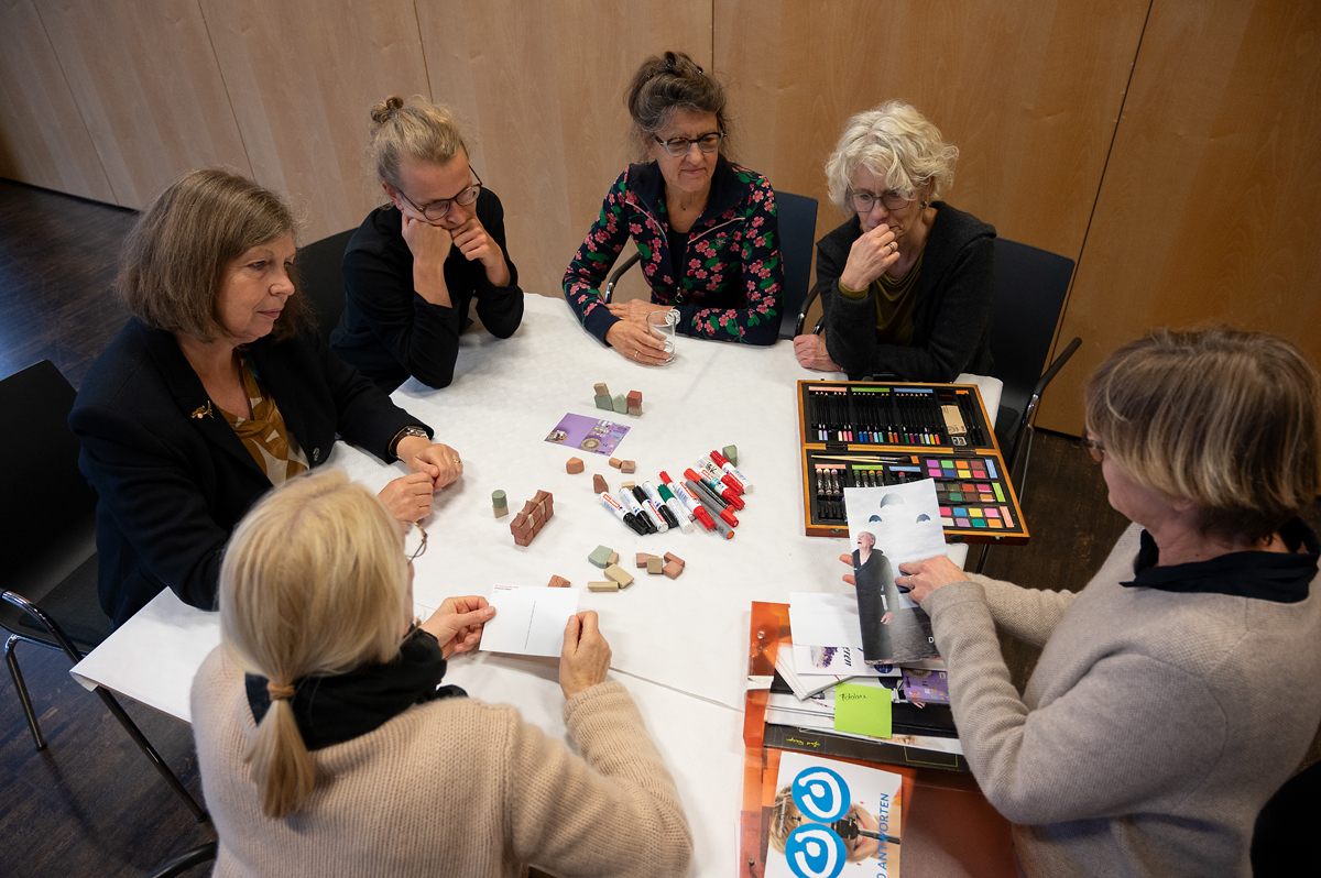 Mehrere Personen sitzen um einen weißen Tisch. Auf dem Tisch liegen Stifte, kleine Spielbausteine, ein Kasten mit Künstlerfarben und verschiedene Flyer