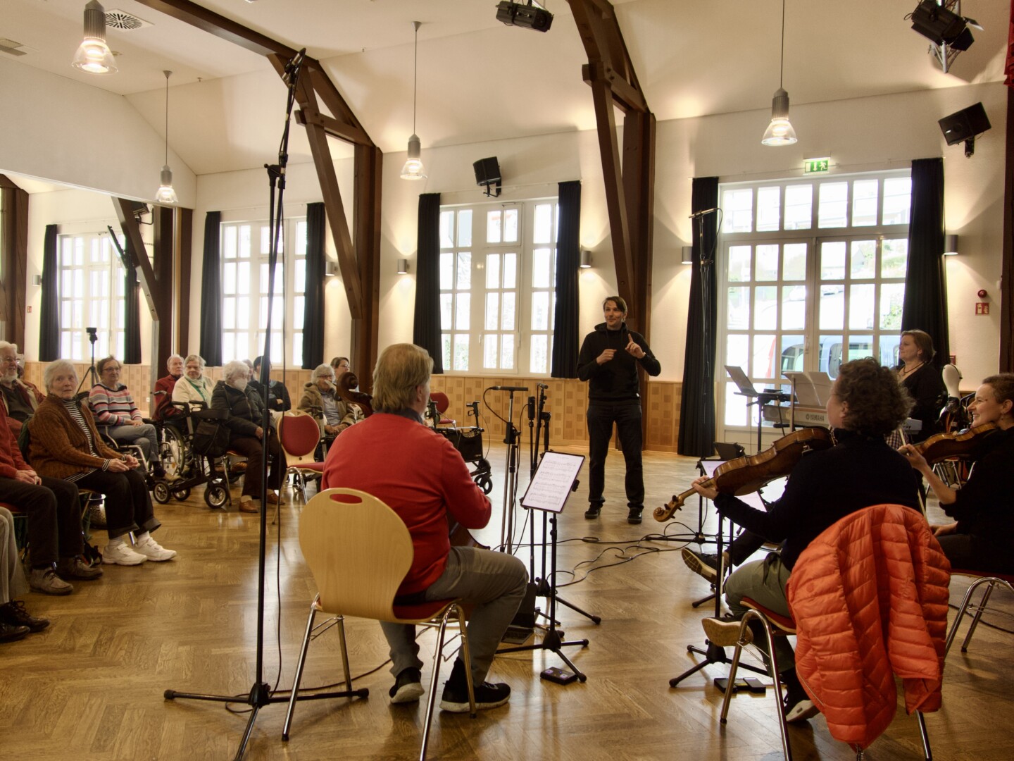 Vier Personen sitzen und spielen ein Instrument, während andere Personen dabei zuschauen.