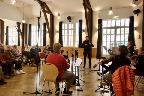 Vier Personen sitzen und spielen ein Instrument, während andere Personen dabei zuschauen.