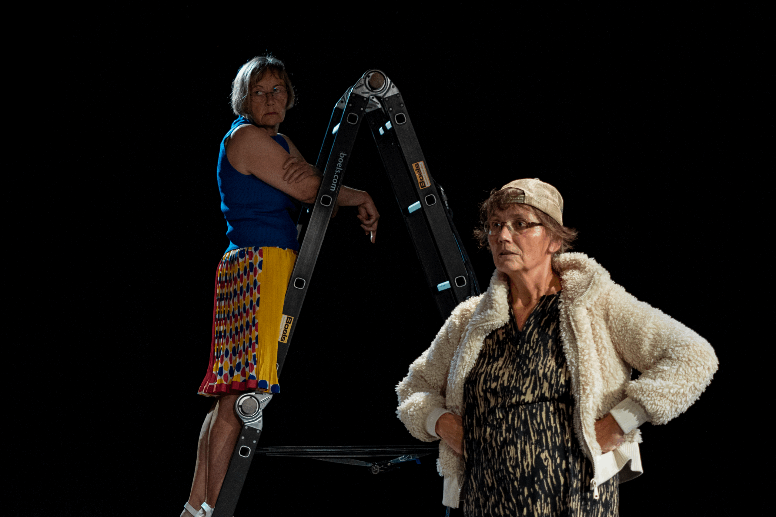 Eine Frau steht mit ihren Händen an der Hüfte im Vordergrund. Im Hintergrund steht eine Frau auf einer Leiter.