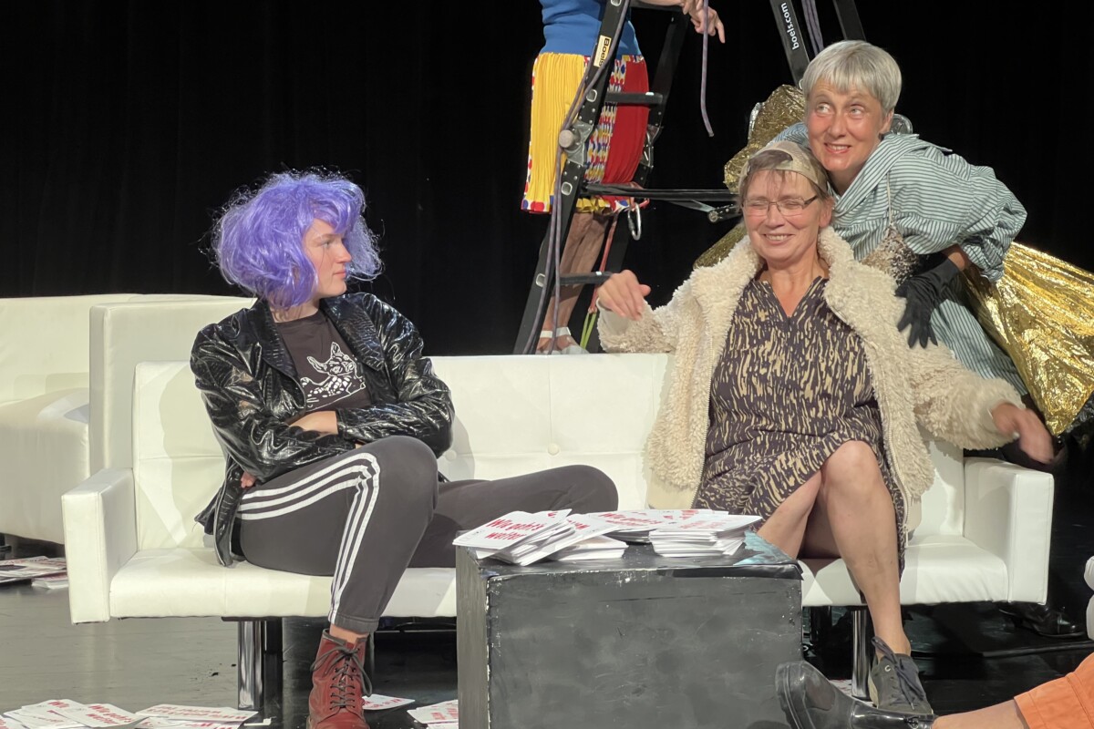 Auf einem Sofa sitzen zwei Frauen. Eine weitere Frau lehnt sich von hinten über die Schulter einer der Frauen.