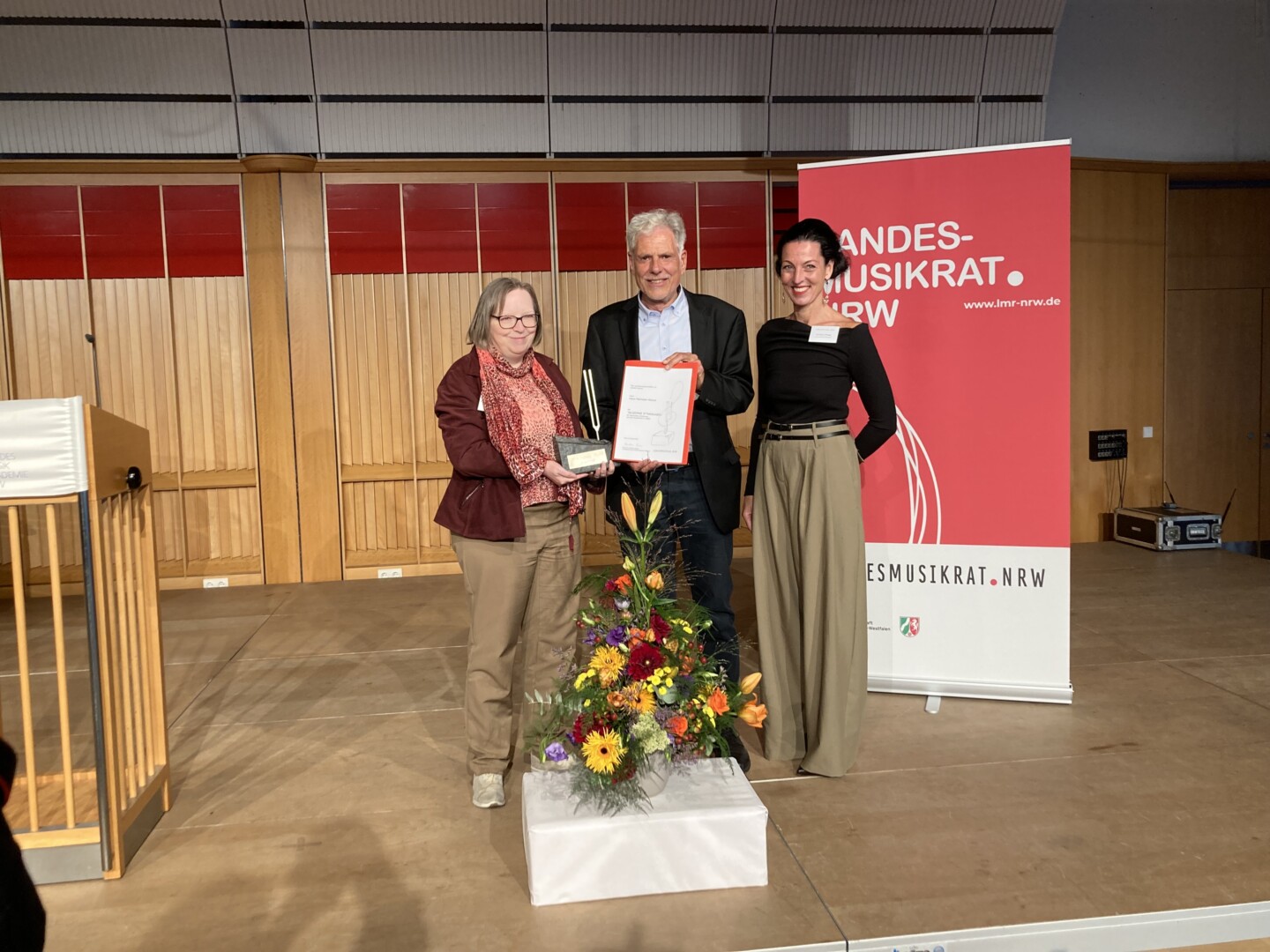 Zwei Frauen stehen links und rechts neben einem Mann auf einer Bühne, der eine Auszeichnung entgegennimmt. Links steht ein Rednerpult, rechts ein Roll-Up mit der Aufschrift „LANDESMUSIKRAT NRW“.