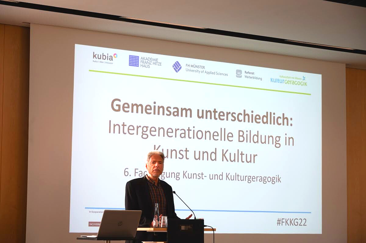 Hans Hermann Wickel, ein älterer Mann mit grauen Haaren, steht an einem Rednerpult. Hinter ihm ist eine Präsentation mit dem Titel „Gemeinsam unterschiedlich: Intergenerationelle Bildung in Kunst und Kultur. 6. Fachtagung Kunst- und Kulturgeragogik“ zu sehen.