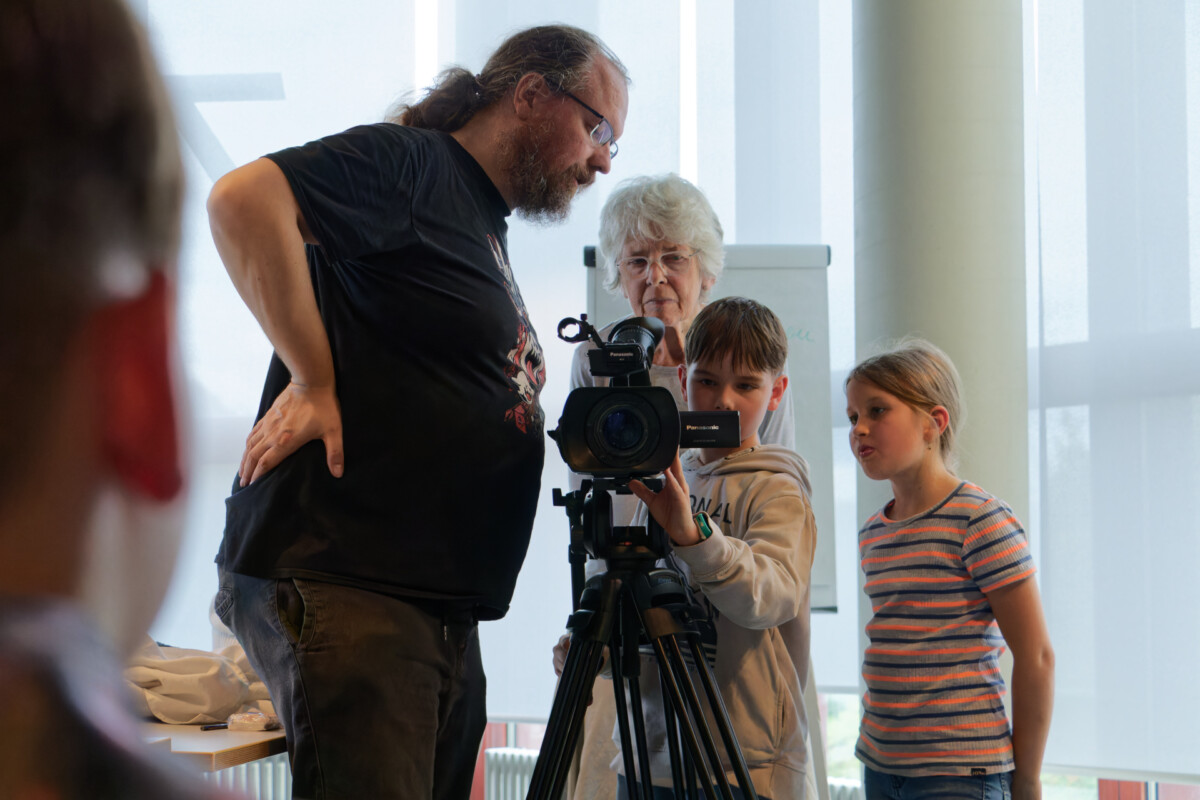 Ein Mann, eine ältere Frau und zwei Kinder stehen an einer Videokamera auf Stativ. Das Kind vorne bedient die Kamera, die anderen schauen zu.