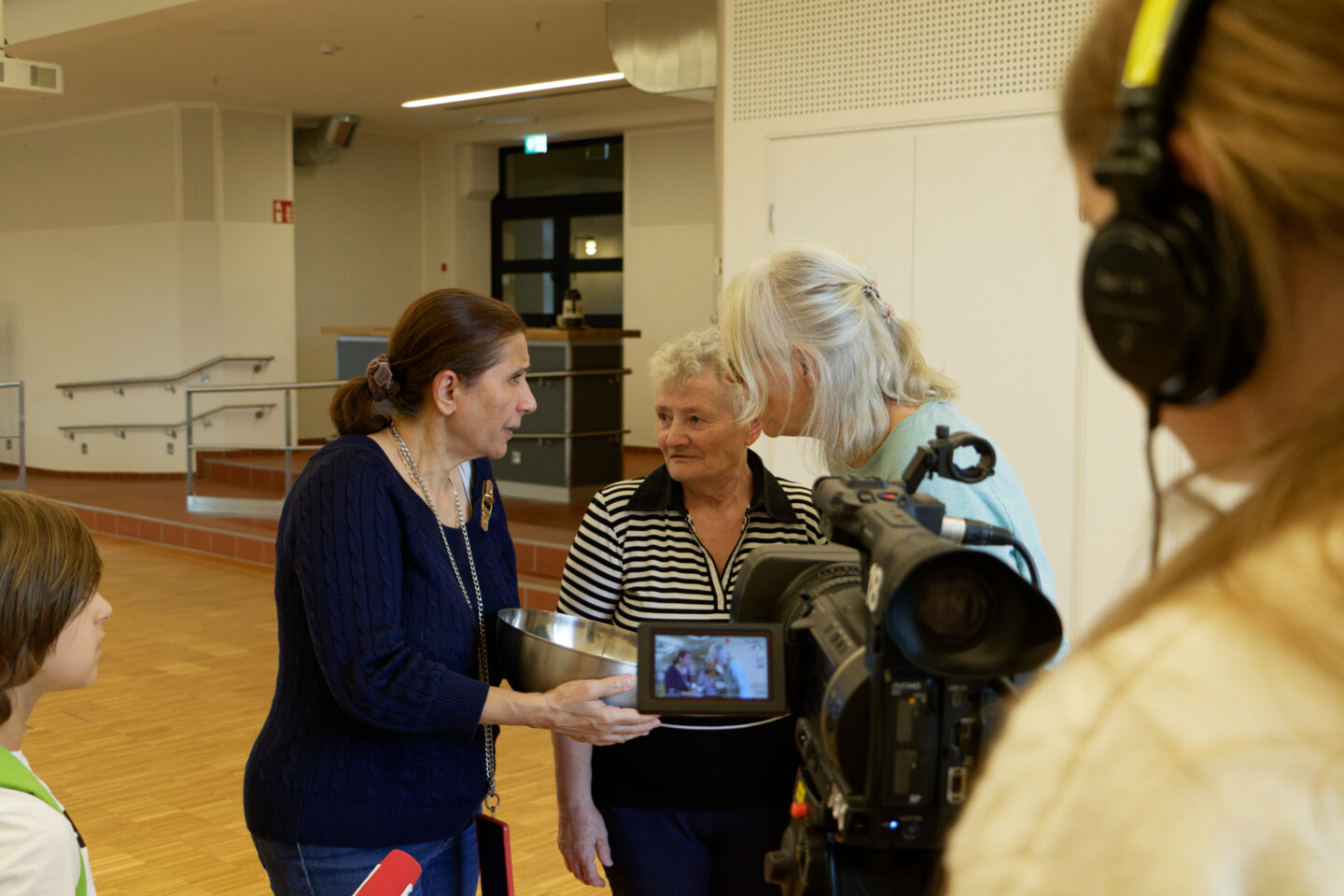 Eine Frau hält eine Metallschüssel und spricht mit zwei älteren Frauen. Eine Kamera filmt die Szene, daneben steht eine Person mit Kopfhörern. Ein Kind beobachtet die Situation von der Seite.
