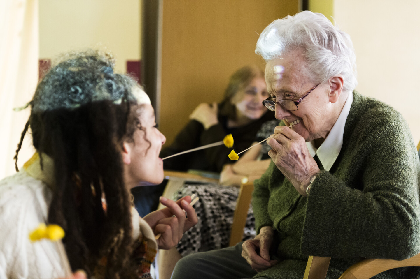 An older woman and a younger woman sit opposite each other, holding two yellow flowers or similar objects in their mouths, which are attached to thin sticks. In the background, other people sit and watch the scene, which is part of an interactive theatre performance.