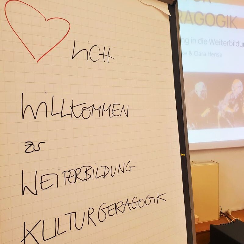 Ein Flipchart zeigt die handschriftliche Begrüßung: „♥ lich Willkommen zur Weiterbildung Kulturgeragogik“.