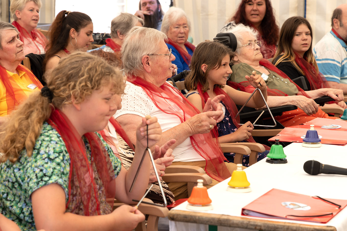 Fotografie: Ältere und jüngere Personen sowie Kinder sitzen in Reihen an Tischen. Einige schlagen Triangeln oder kleine Glocken an.