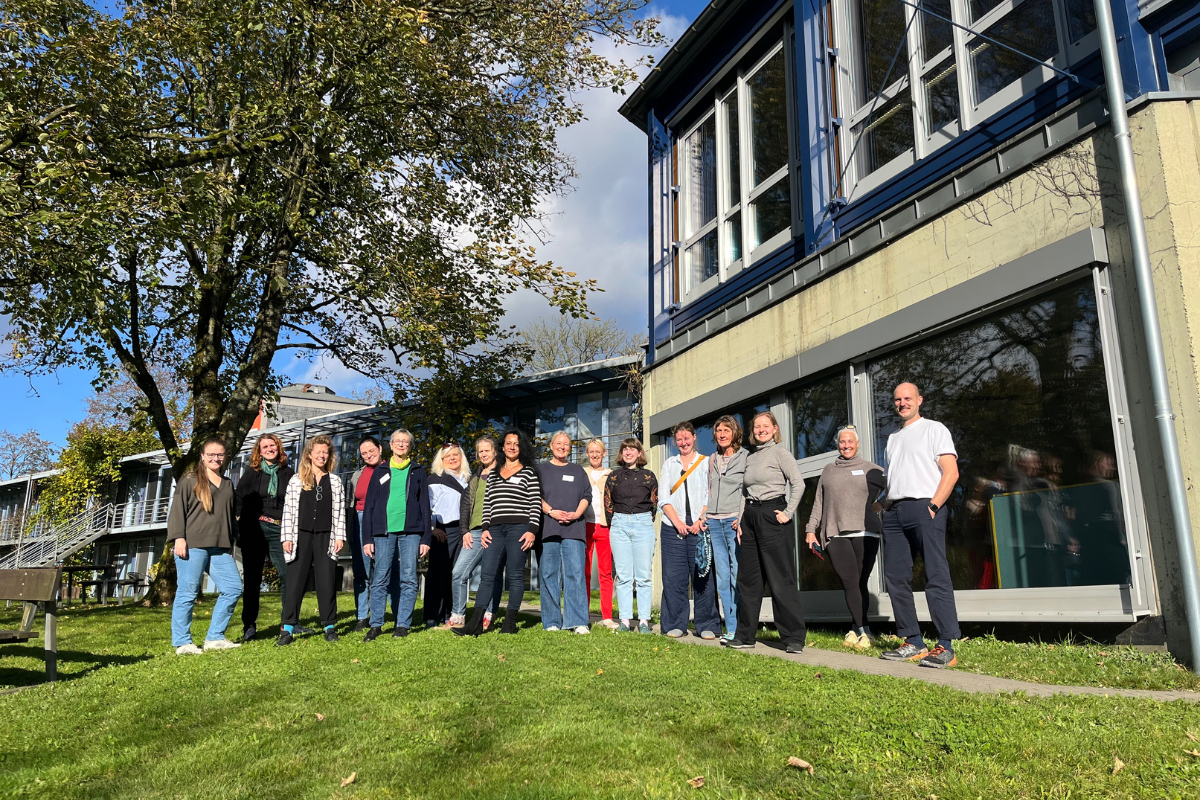Eine Gruppe von 15 Personen steht auf einer Wiese vor einem modernen Gebäude mit großen Fenstern. Links steht ein Baum, im Hintergrund sind weitere Gebäudeteile sichtbar.