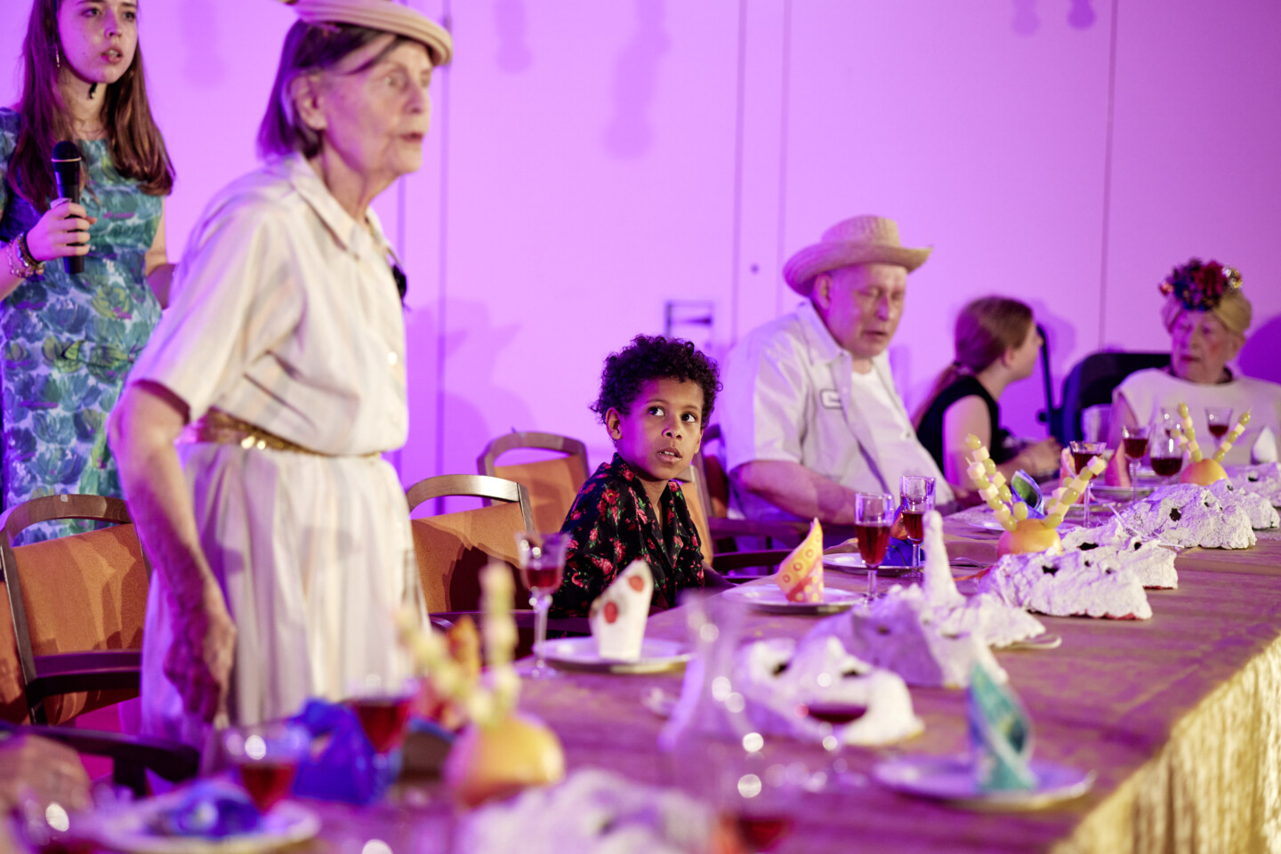 Fotografie: Ältere Personen und Kinder sitzen und stehen an einem festlich gedeckten Tisch. Vor ihnen liegen Gipsmasken.