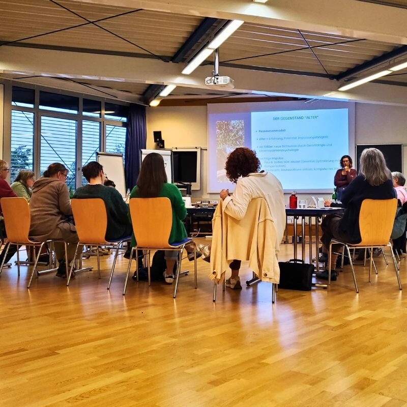 Mehrere Personen sitzen in einem Seminarraum an Tischen. Auf einer Leinwand steht eine Präsentation mit dem Titel „Der Gegenstand ‚Alter‘“.