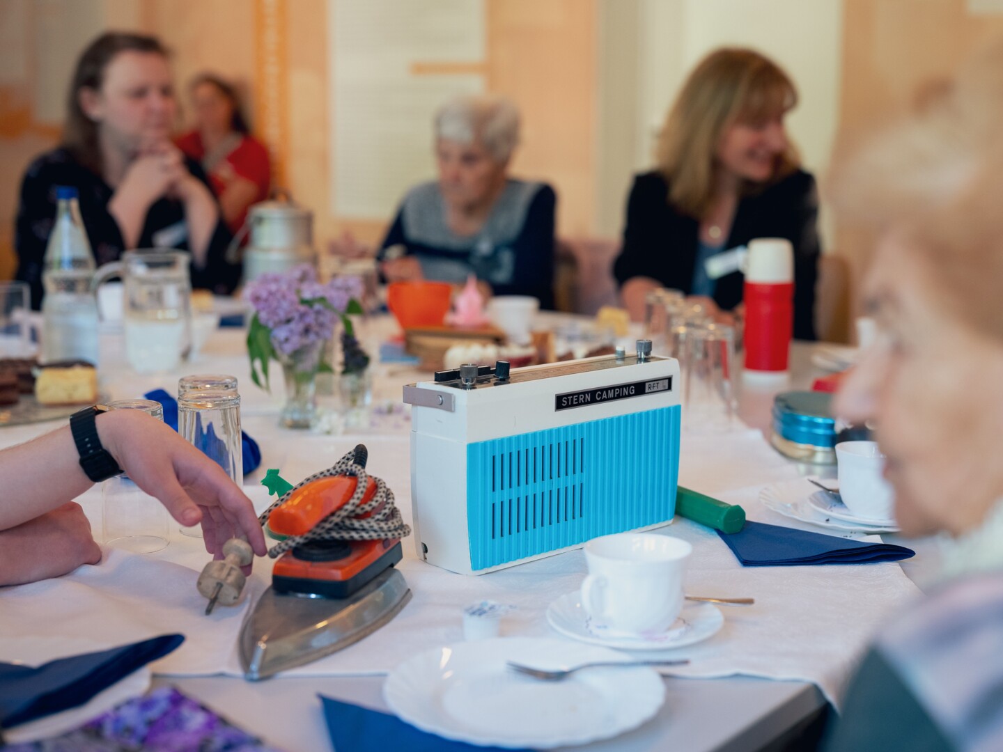 Fotografie: Retro-Radioempfänger und Retro-Bügeleisen auf einem gedeckten Tisch mit Kaffeetasse. Mehrere Personen unterschiedlichen Alters sitzen am Tisch.
