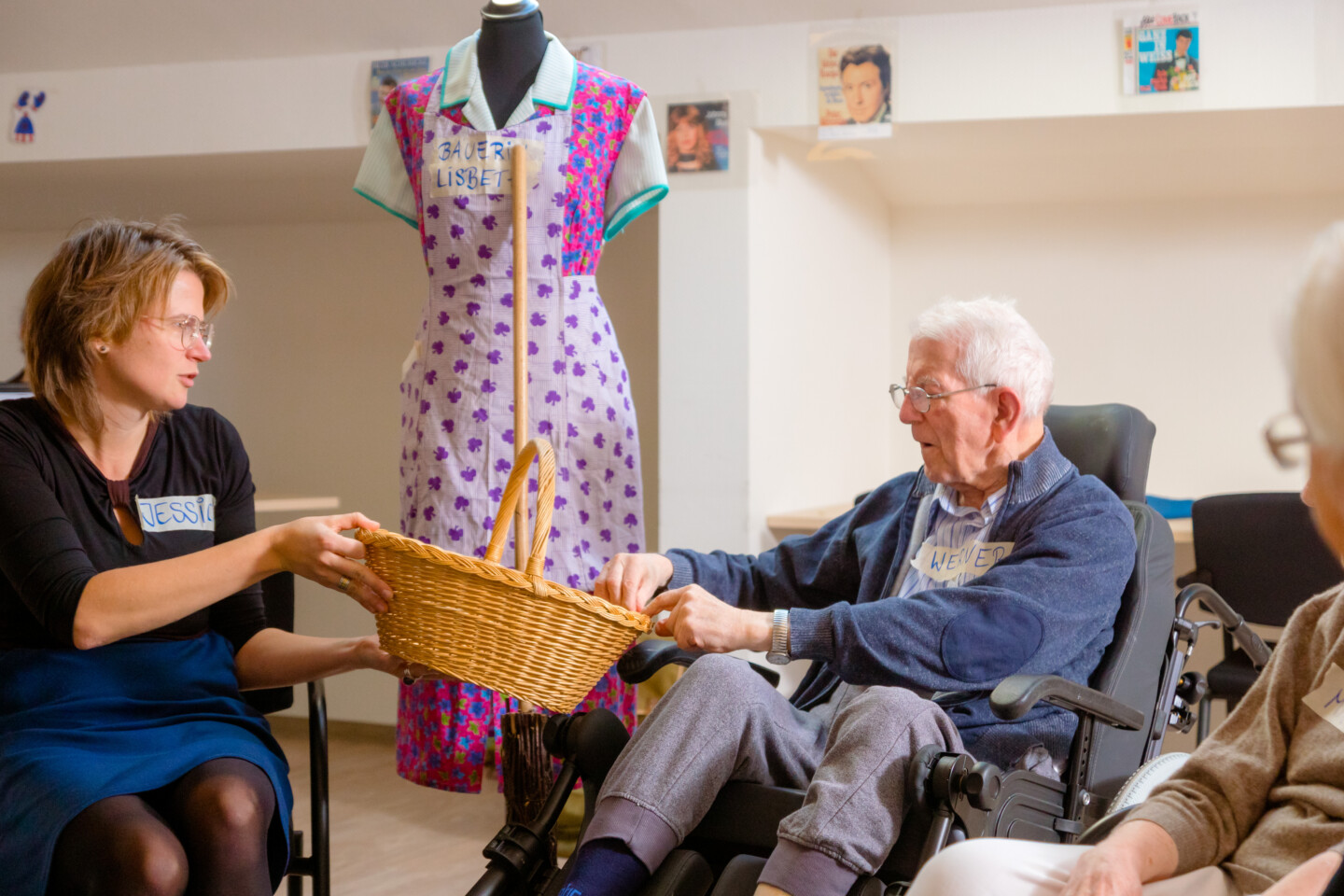 Fotografie: Eine jüngere Frau und ein älterer Mann im Rollstuhl reichen sich einen geflochtenen Korb. Im Hintergrund steht eine Schaufensterpuppe in gemustertem Kleid und Kittelschürze.
