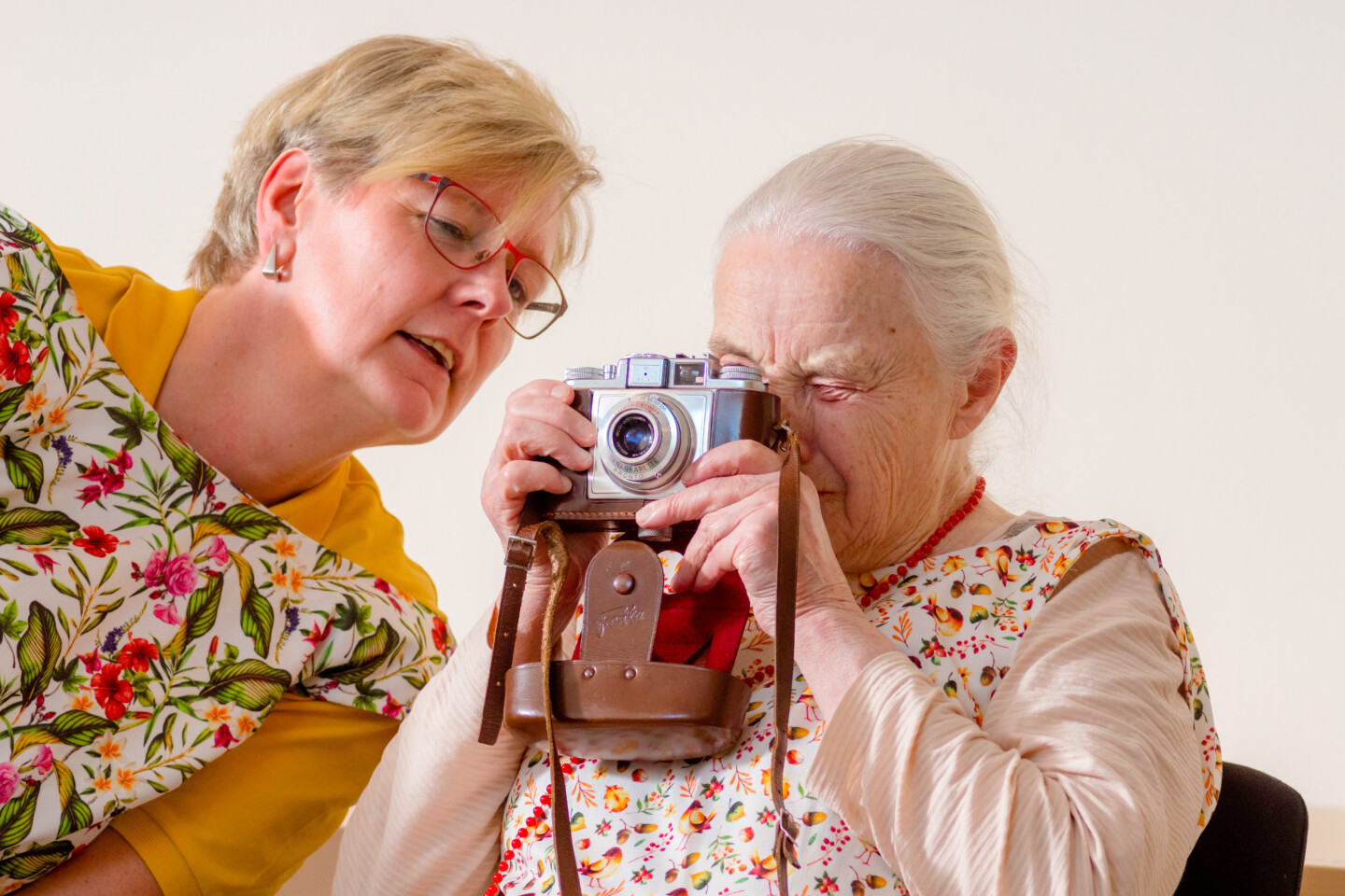 Fotografie: Eine ältere Frau und eine mittleren Alters, beide mit gemusterten Schürzen, betrachten gemeinsam eine analoge Kamera mit Ledertasche.