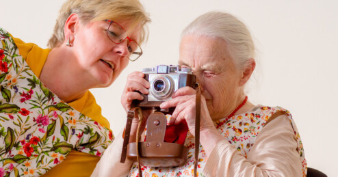 Fotografie: Eine ältere Frau und eine mittleren Alters, beide mit gemusterten Schürzen, betrachten gemeinsam eine analoge Kamera mit Ledertasche.