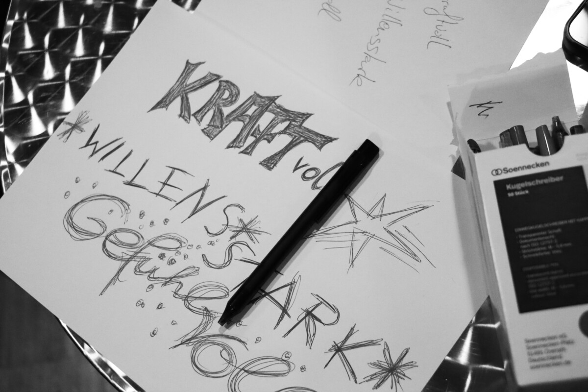 Ein mit handgezeichneten Worten „kraftvoll“, „willensstark“ und „gefühlsvoll“, verziert mit Sternen. Daneben liegen ein Stift und eine Schachtel Kugelschreiber.
