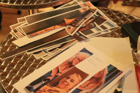 Auf einem runden Metalltisch liegen in Streifen zerschnittene Fotos einer älteren Frau. Unter den Streifen liegt ein Blatt Papier, auf dem handschriftlich steht: „so fröhlich und neugierig“.