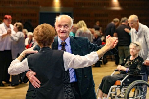 Ein älterer Mann und eine Frau tanzen. Im Hintergrund sind tanzende Paare und eine alte Frau im Rollstuhl zu sehen.