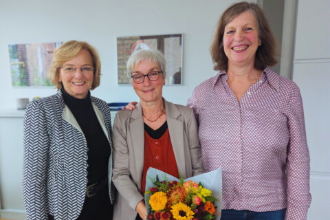 Brigitte Schorn, Gerda Sieben und Almuth Fricke stehen lächelnd nebeneinander in einem hellen Raum. Gerda Sieben hält einen orange-gelben Blumenstrauß in den Händen.