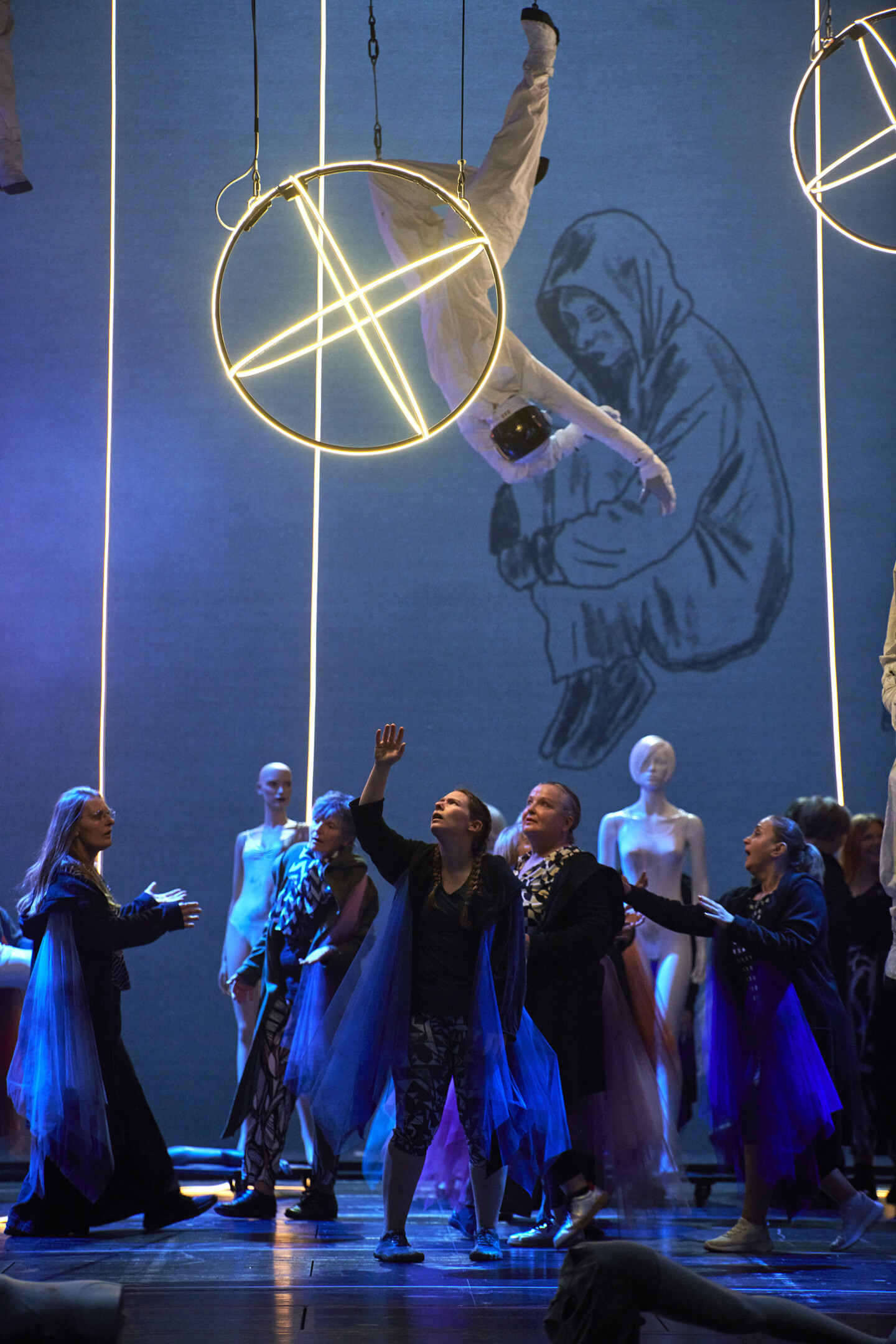 Fotografie: Eine Person in weißem Anzug hängt kopfüber hinter einem beleuchteten Metallring. Mehrere Personen stehen darunter und blicken zu ihr auf.