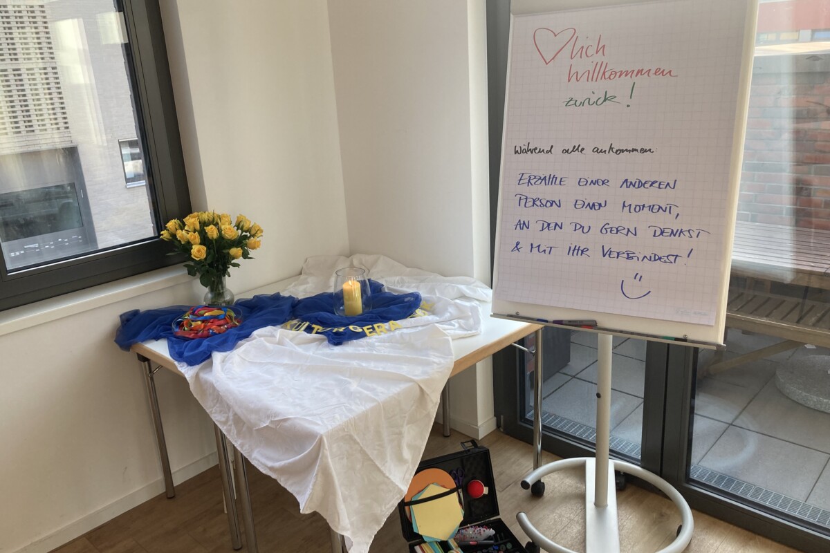 Eindruck aus dem Kursraum. Auf einer Flipchart werden die Teilnehmenden zum letzten Modul begrüßt und können mit den anderen ins Gespräch kommen.