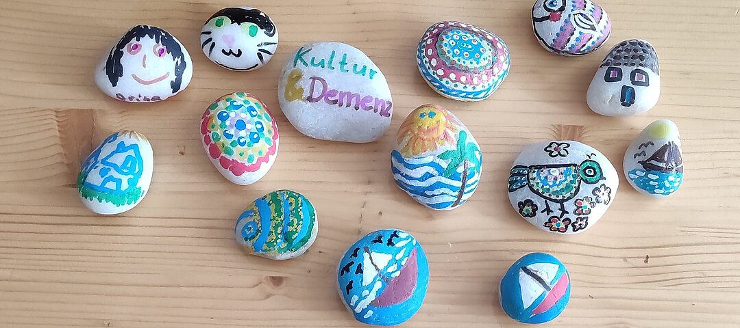 Bunt bemalte Kieselsteine. Auf einem der Steine steht "Kultur & Demenz".