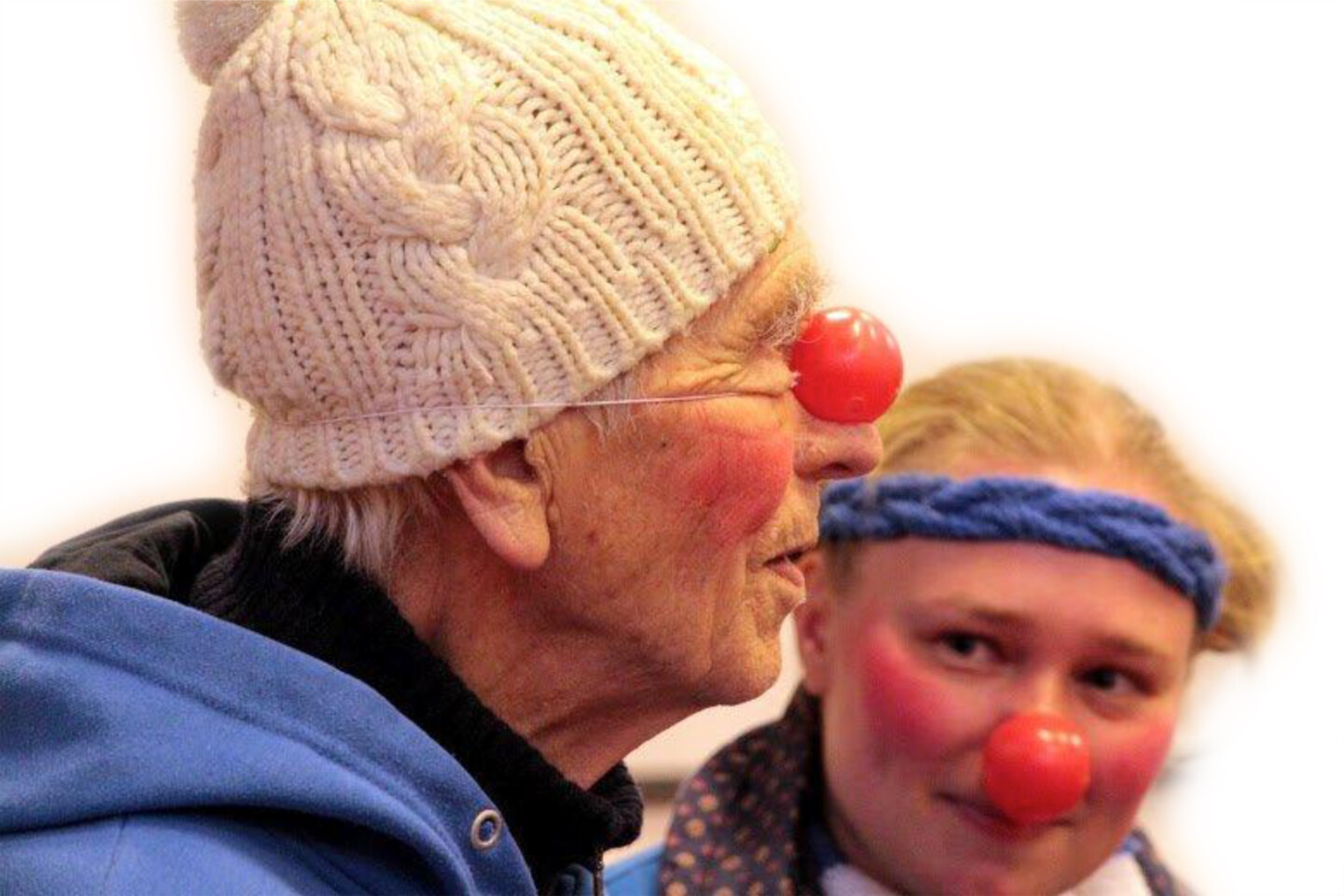 Fotografie: Älterer Mann mit Strickmütze und junge Frau mit Stirnband, beide tragen eine rote Clownsnase.