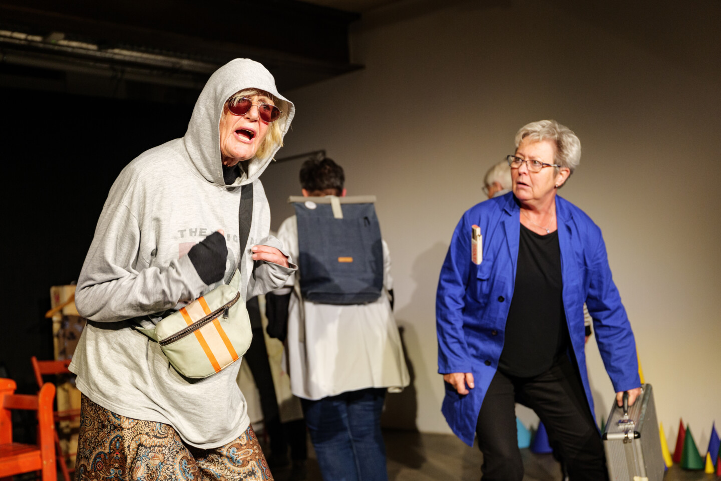 Theaterszene mit zwei älteren Frauen: Eine trägt eine Sonnenbrille, einen grauen Hoodie mit Kapuze über dem Kopf, eine andere einen blauen Arbeitskittel und einen silbernen Koffer.