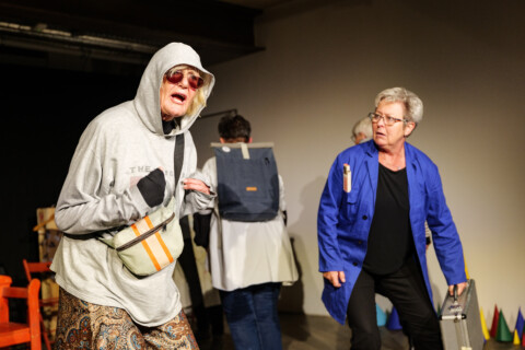 Theaterszene mit zwei älteren Frauen: Eine trägt eine Sonnenbrille, einen grauen Hoodie mit Kapuze über dem Kopf, eine andere einen blauen Arbeitskittel und einen silbernen Koffer.