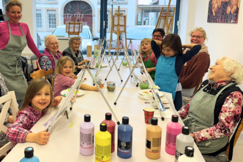 Mehrere ältere Frauen und Kinder sitzen gemeinsam an einem langen Tisch und malen. Vor ihnen stehen kleine Staffeleien mit Leinwänden sowie bunte Farbflaschen. Einige Personen tragen Malschürzen. Im Hintergrund sind große Fenster und weitere Staffeleien zu sehen.