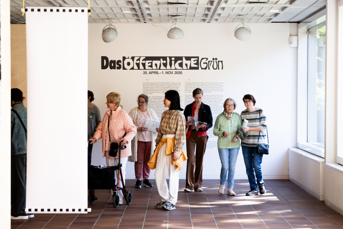 Mehrere ältere Frauen und eine jüngere Frau bewegen sich durch einen Ausstellungsraum. Einige halten Broschüren in den Händen, eine Frau nutzt einen Rollator. An der Wand steht: „Das Öffentliche Grün. 25. APRIL–1. NOV. 2026“.